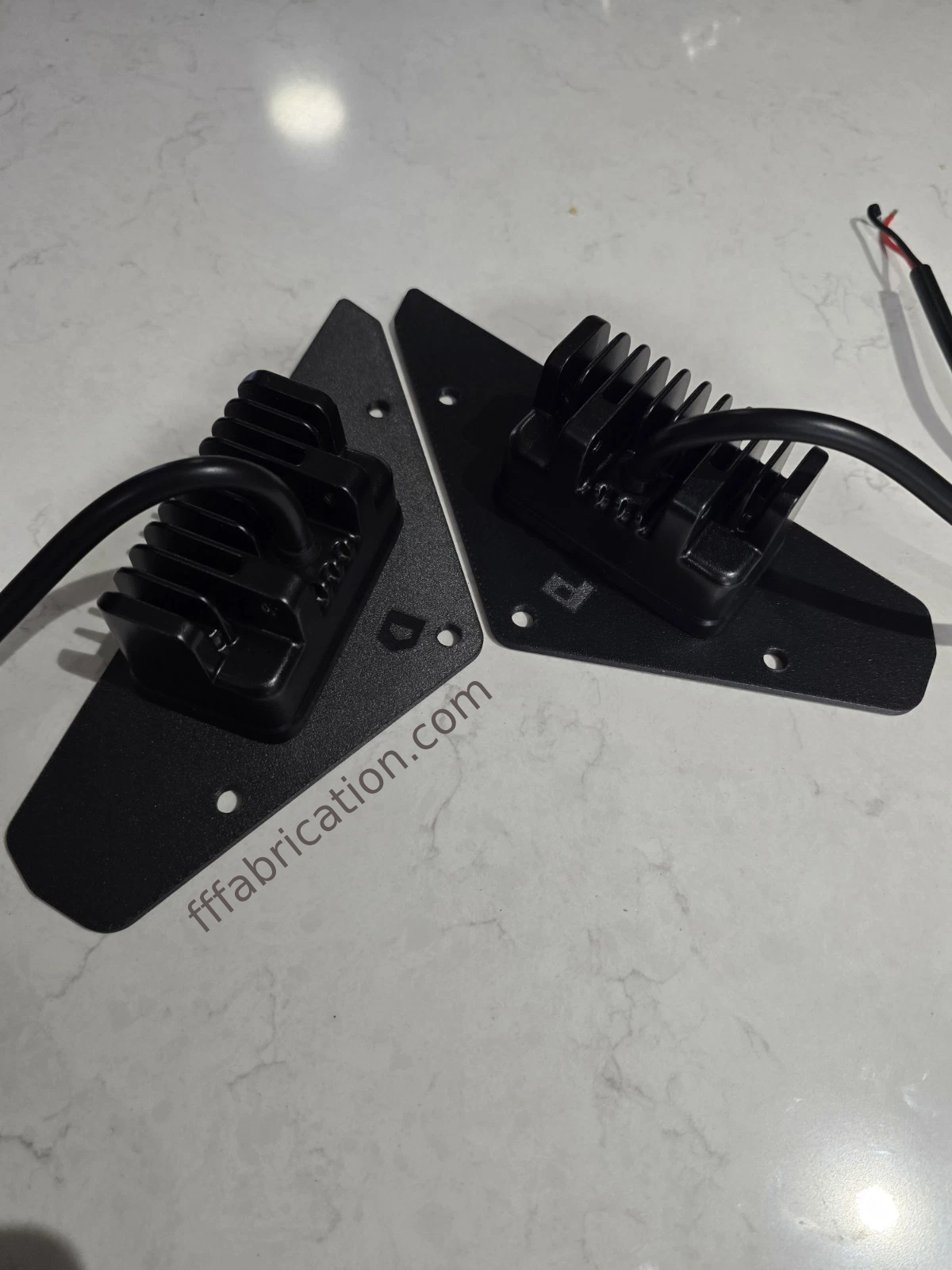 Two black metal brackets with wiring on a white surface, branded 'fffabrication.com'.