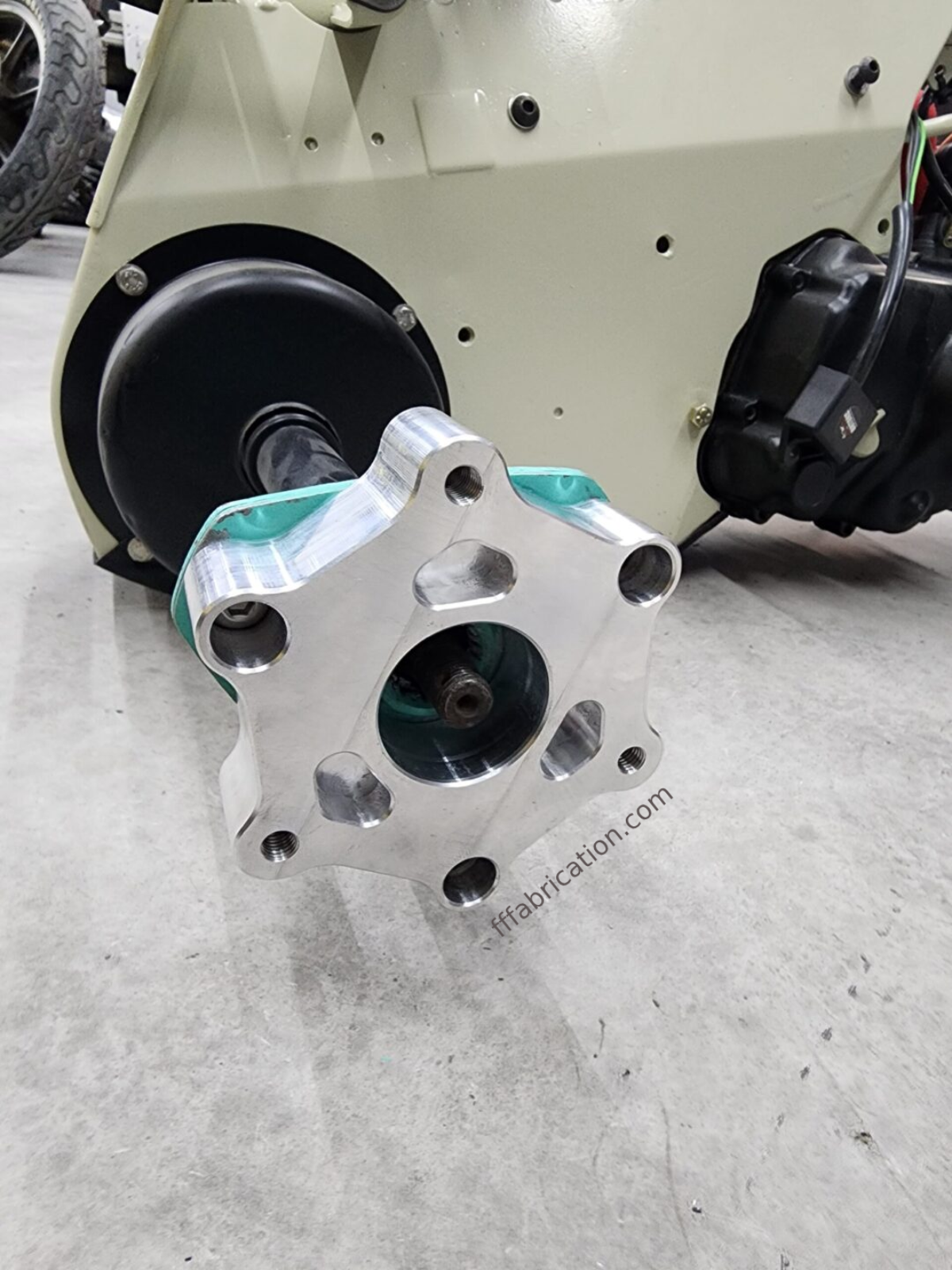 Metallic mechanical component on a concrete floor with 'fabrication.com' branding.